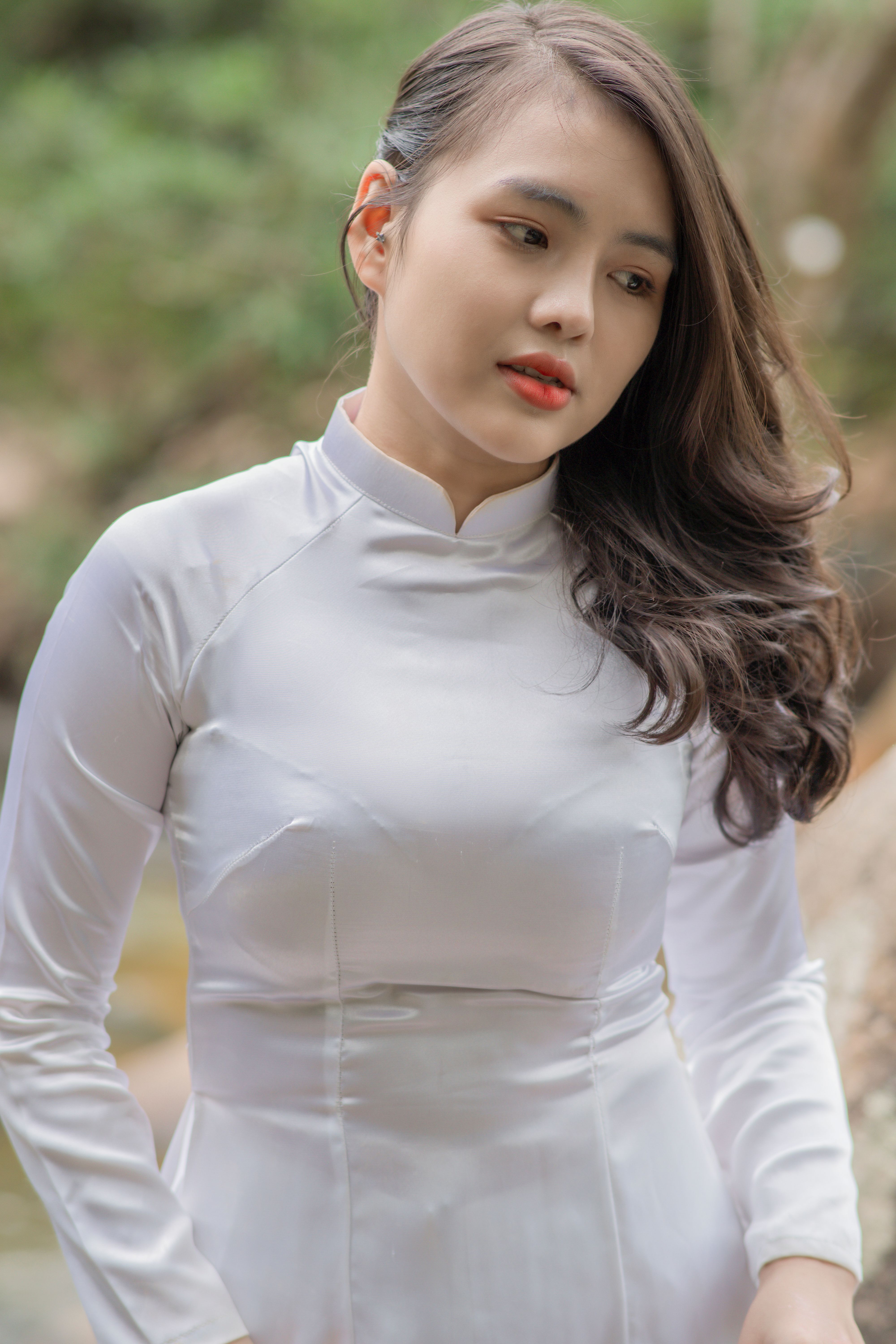 Portrait of a young woman wearing a white traditional dress standing outdoors, with lush greenery behind her.