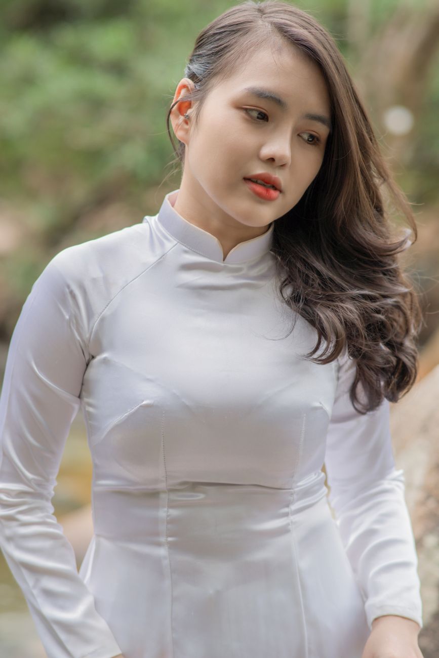 Portrait of a young woman wearing a white traditional dress standing outdoors, with lush greenery behind her.