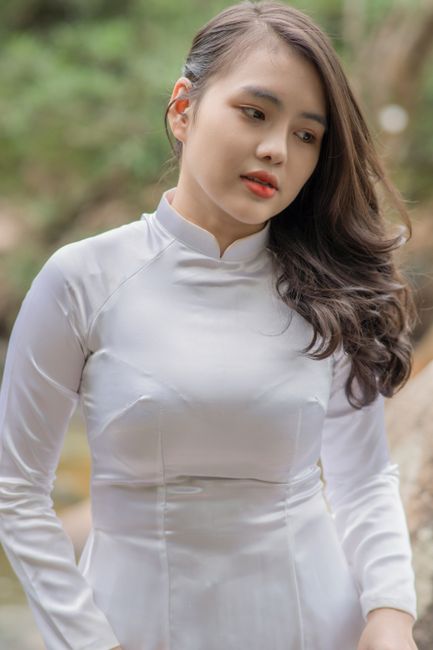 Portrait of a young woman wearing a white traditional dress standing outdoors, with lush greenery behind her.