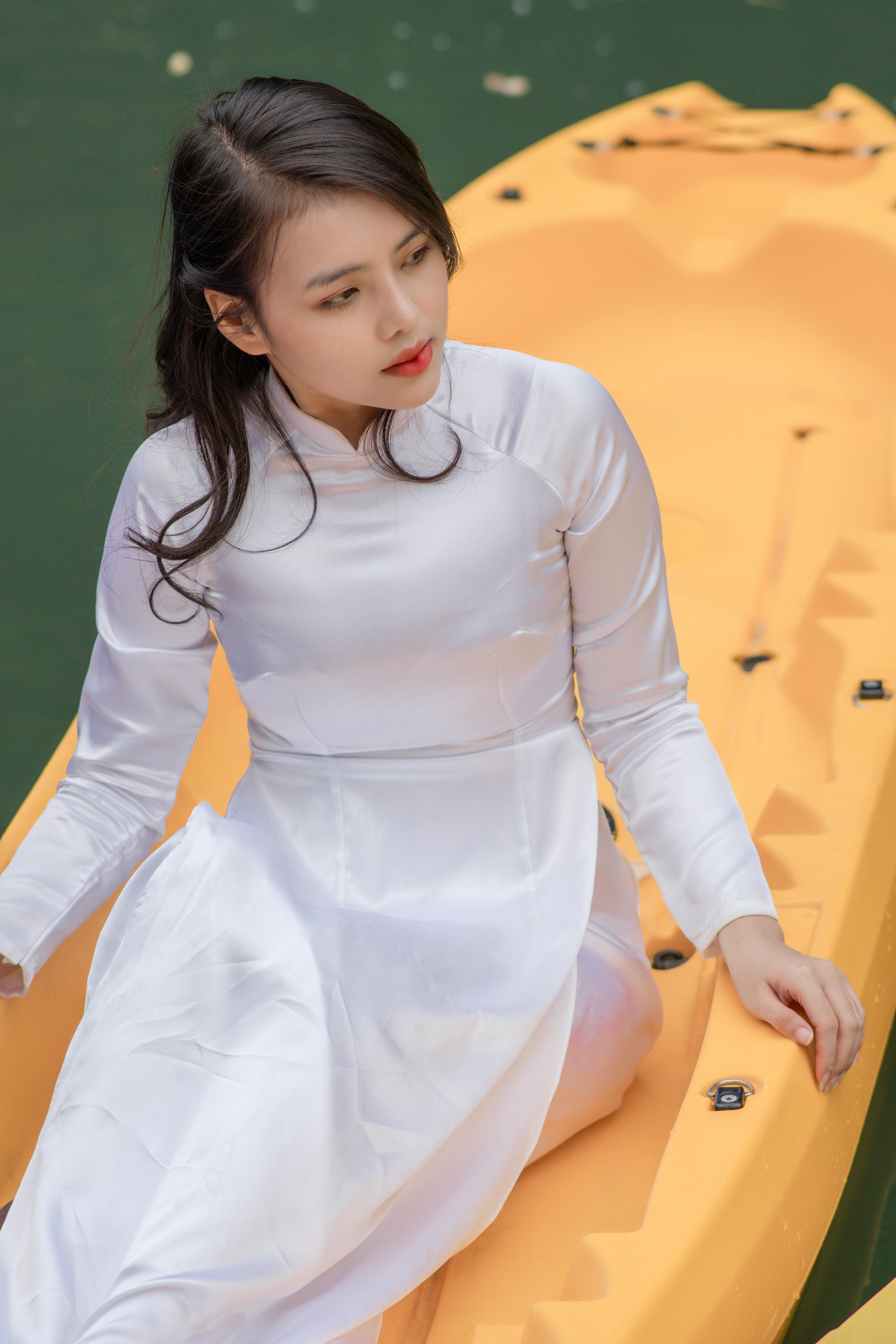A young woman in a white traditional dress sits on a yellow kayak in a serene setting.