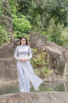 Serene portrait of a woman wearing a traditional white dress standing by a rock pool in nature.