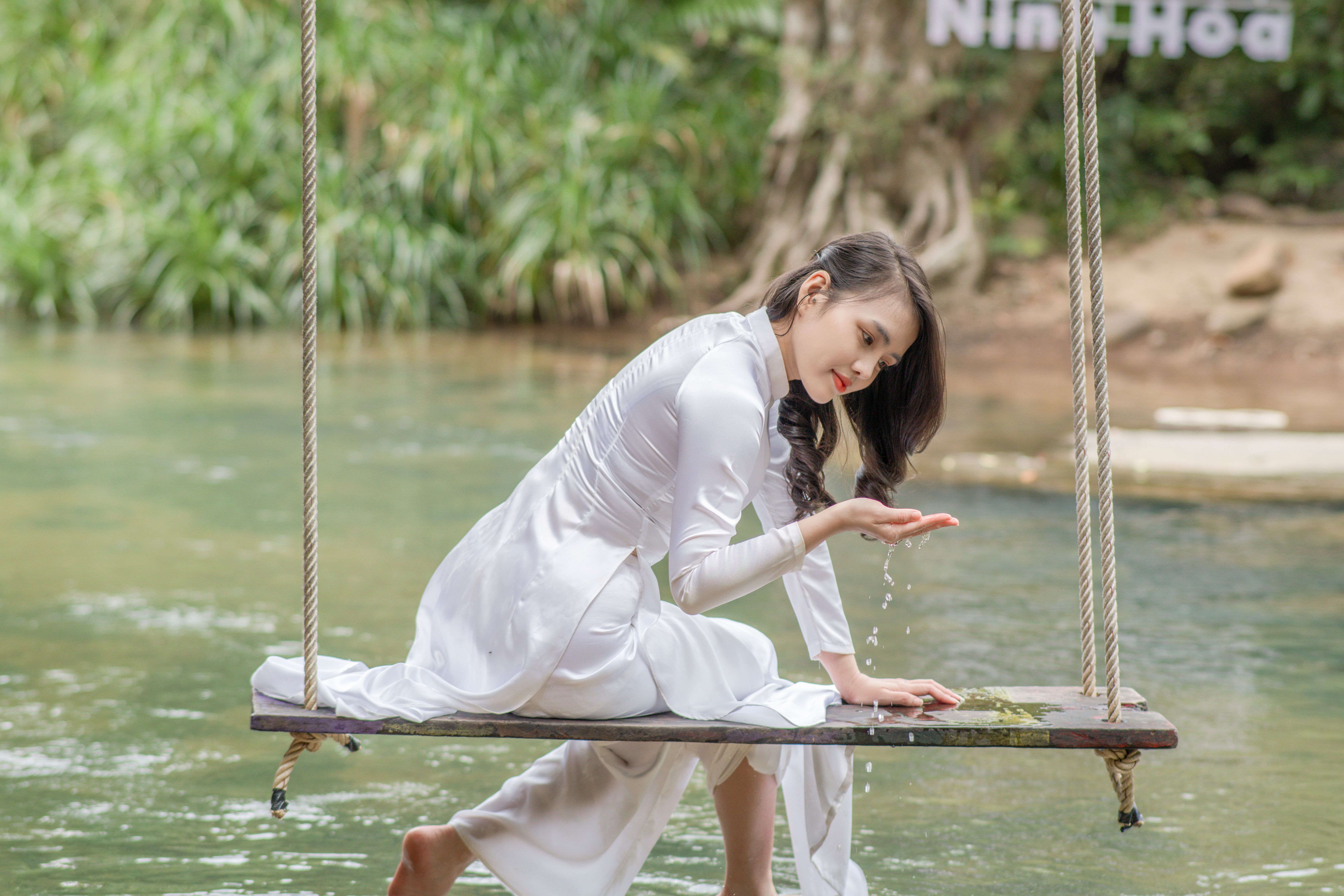 Elegant young woman in traditional attire sits on a river swing, embracing nature.