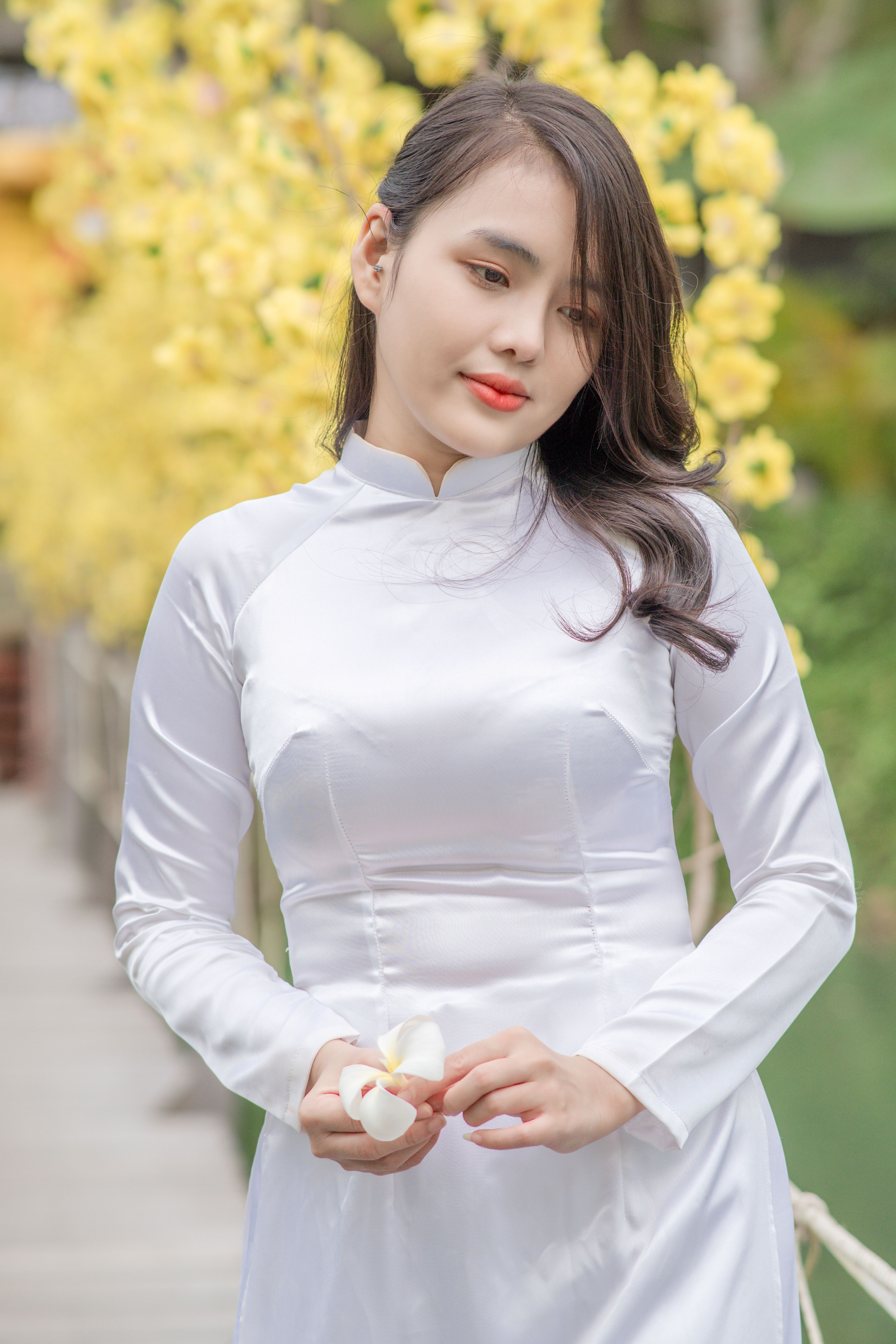 Elegant woman in traditional white Áo Dài amidst yellow blossoms outdoors.