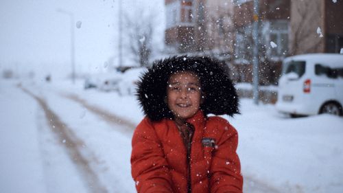 Free A smiling child wearing a red coat enjoys a snowy day in a city street, capturing joyful winter fun. Stock Photo