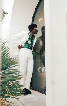 Elegant man in white suit and green shirt with sunglasses standing near a modern glass door.