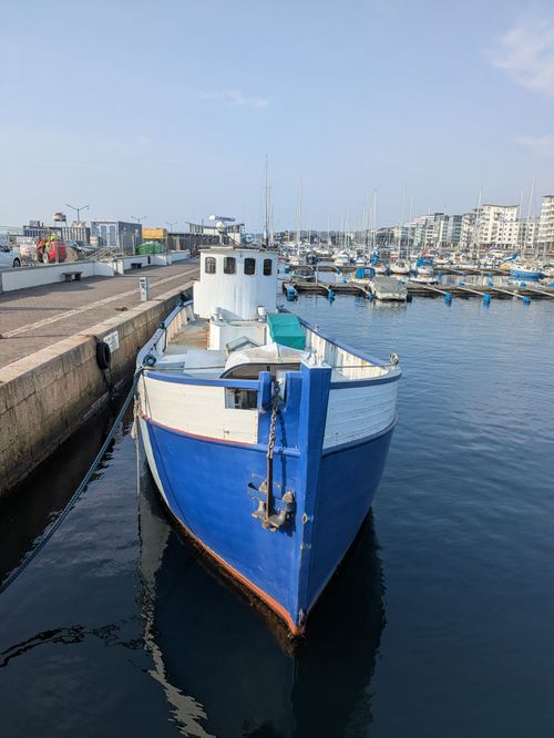Free A classic blue fishing boat docked at a city marina surrounded by modern buildings under a clear sky. Stock Photo
