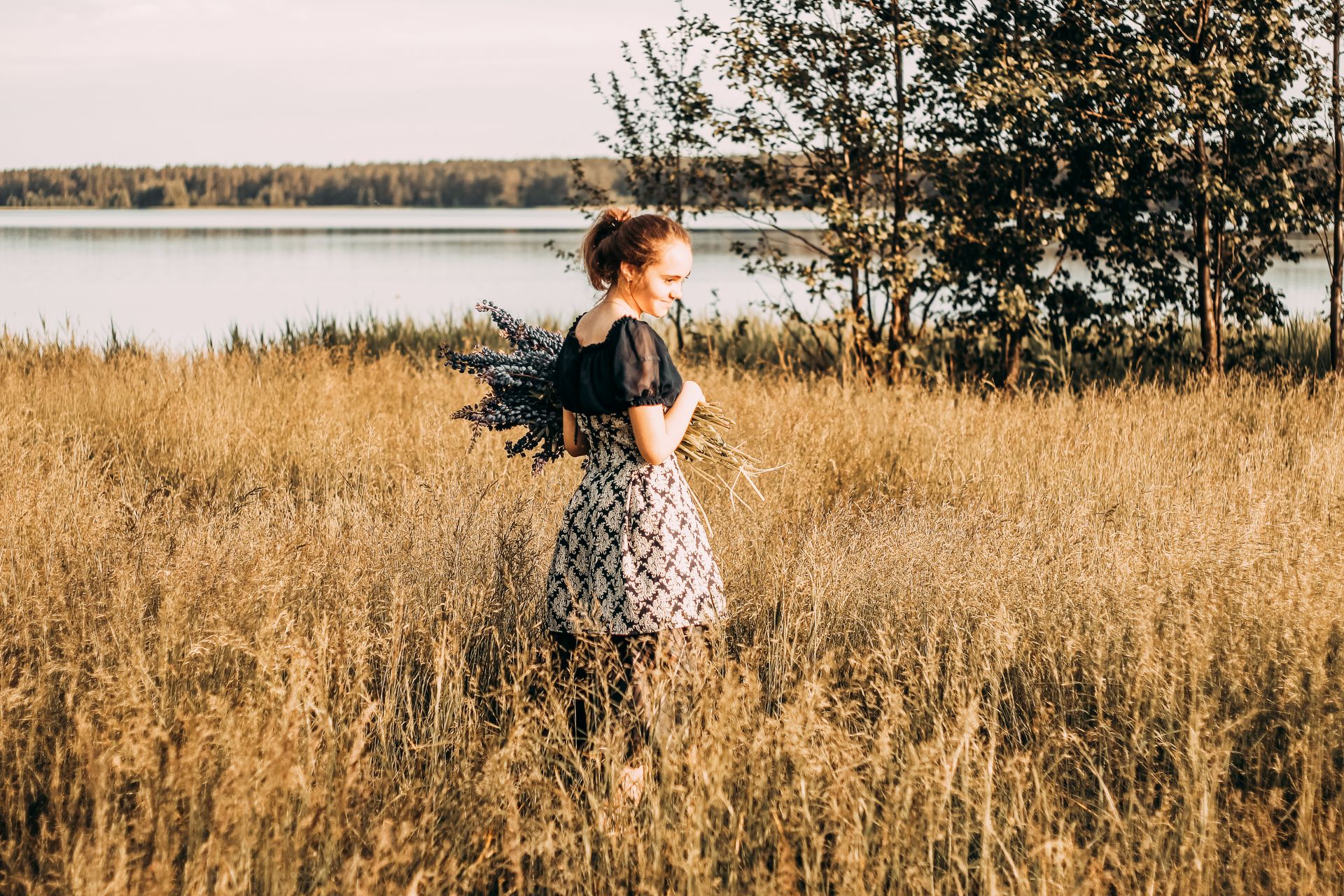 A young woman stands in a sunlit field by a lake, holding lavender flowers and smiling.