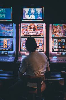Adult male seated in a dimly lit casino engaging with vibrant slot machines at night.