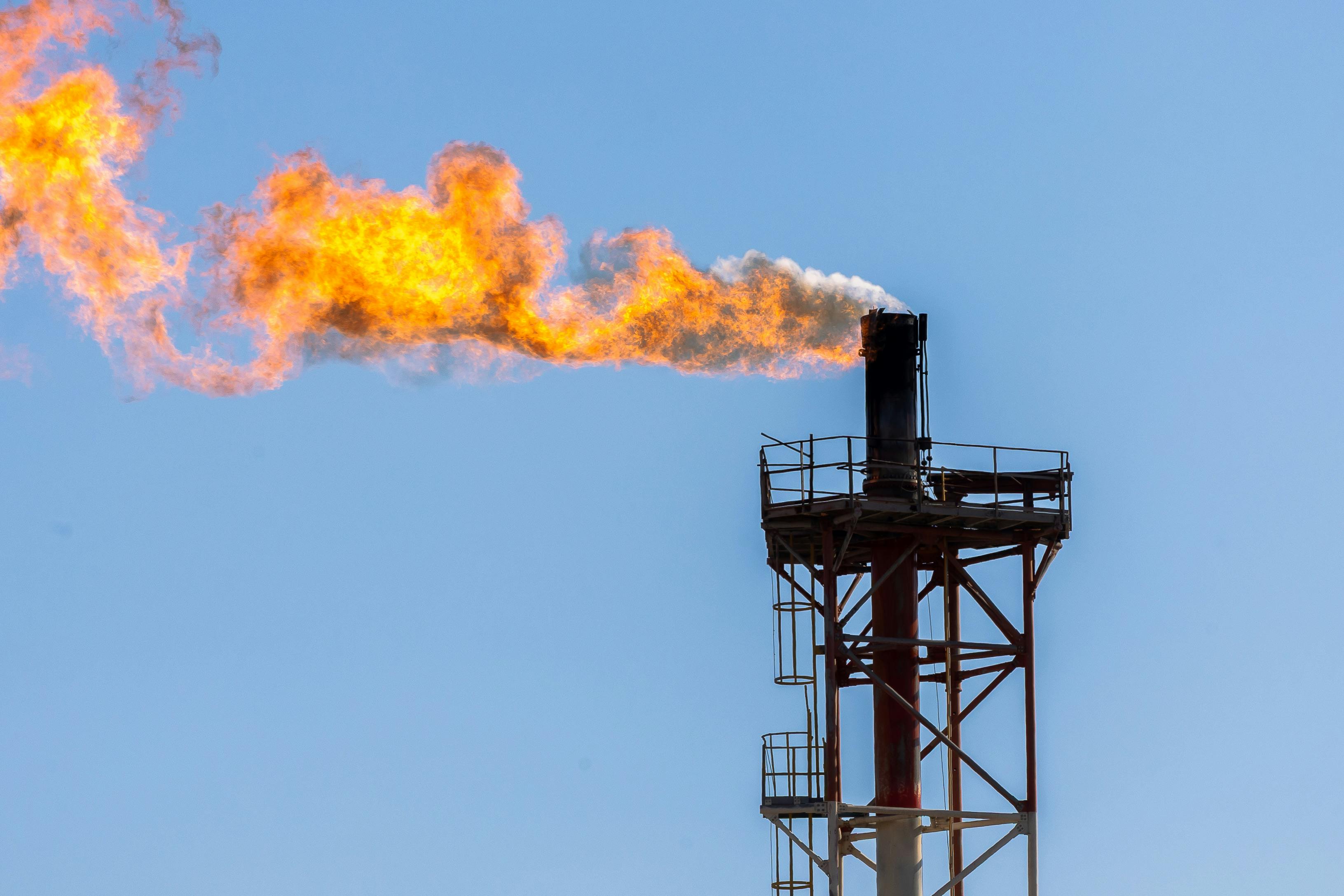 Close-up of an industrial gas flare with a vibrant orange flame against a clear blue sky.