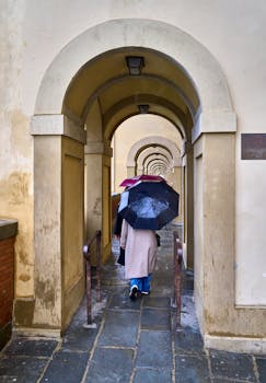 A group of adults with umbrellas walks through ancient stone arches on a rainy day.