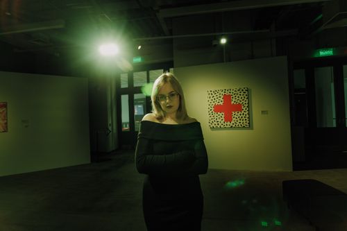 Free A woman in a gallery, illuminated by green light, stands near a red cross art piece. Stock Photo