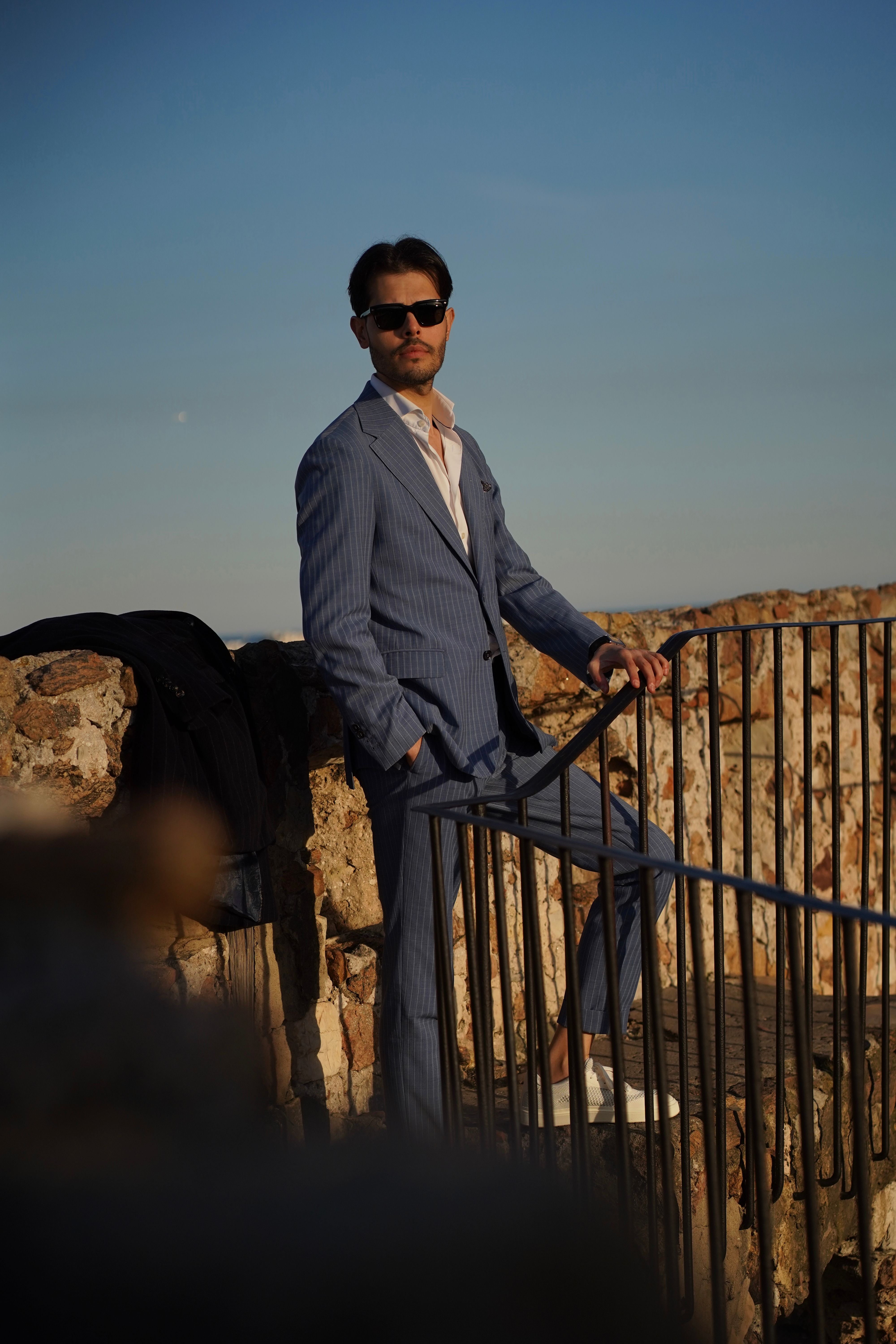 A fashionable male in a suit poses confidently on a historic stone terrace against a blue sky.
