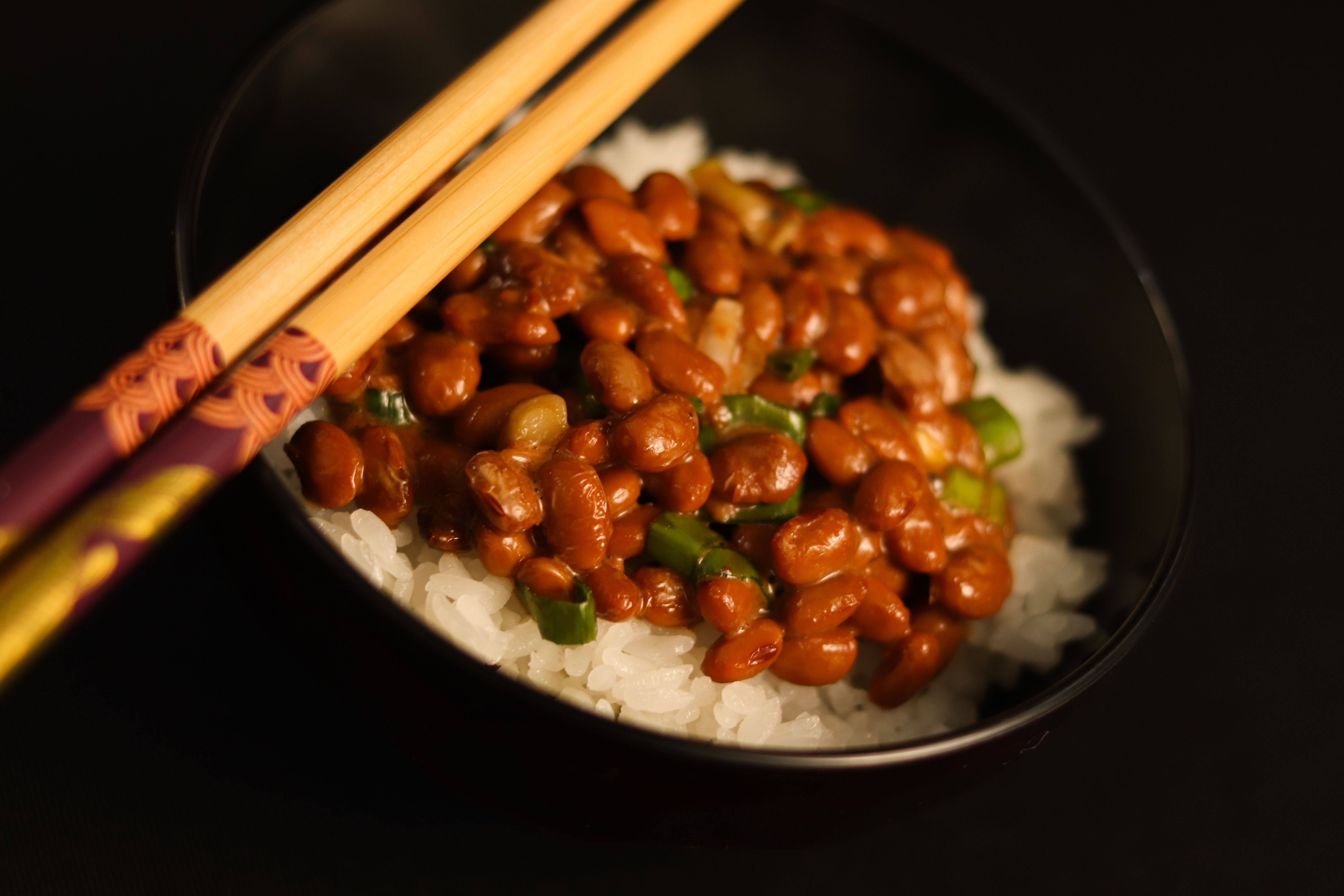 A bowl of natto beans served over white rice with chopsticks on a dark background.