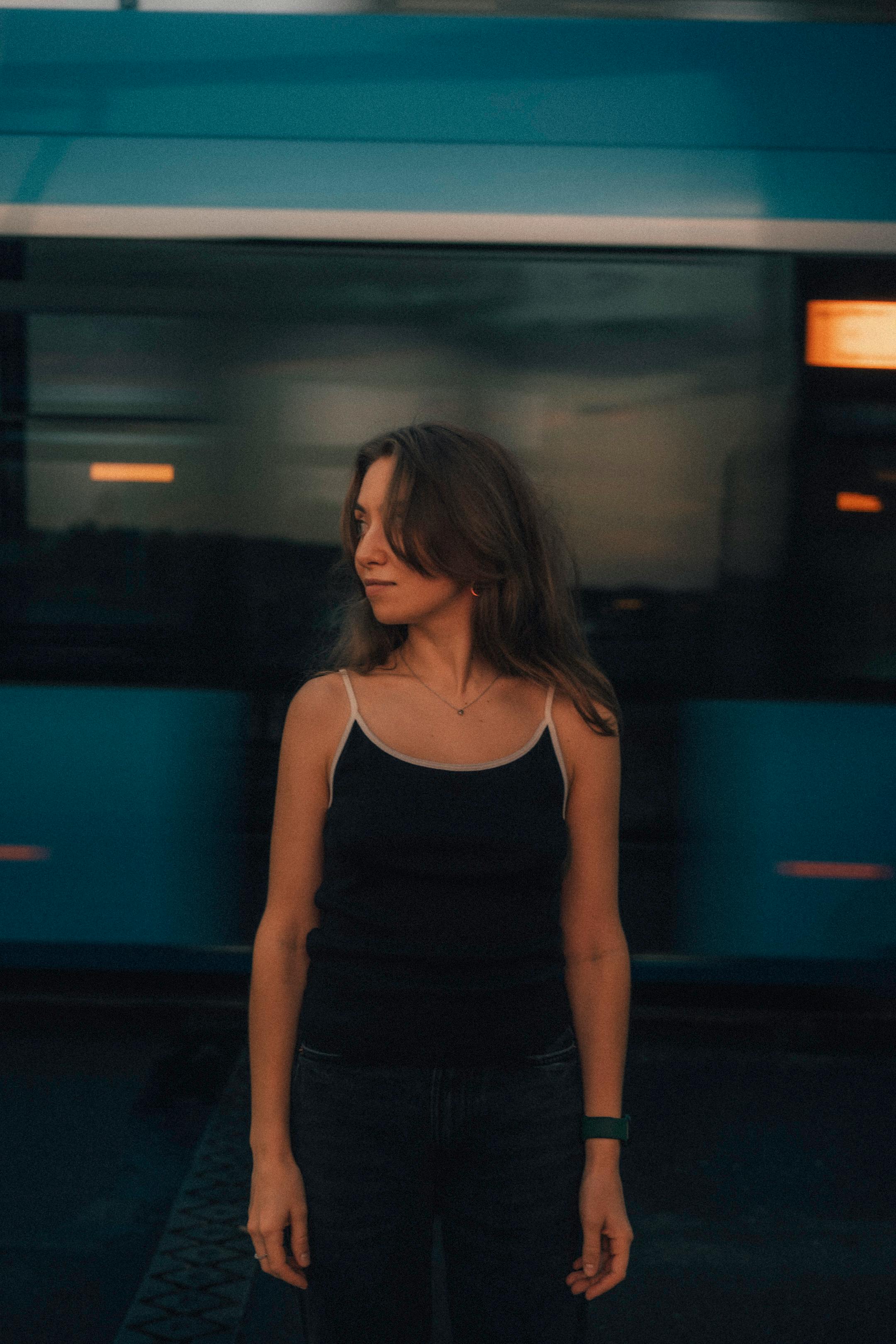 Woman in dark attire stands still as train rushes by at dusk, creating motion blur effect.