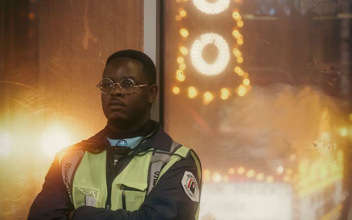 A security guard stands by a neon sign at night, emitting a warm glow.