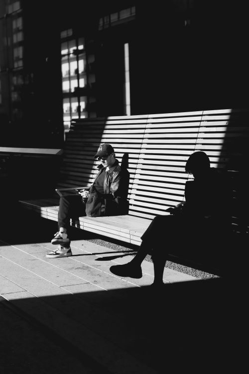 Free Two people sit in shadow and sunlight on a city bench. Black and white contrast emphasizes urban solitude. Stock Photo
