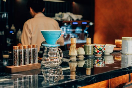 Barista making drip coffee in a cafe with various coffee and tea tools displayed on the counter.