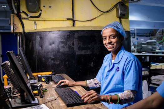 A smiling female factory worker operates a computer in an industrial setting.