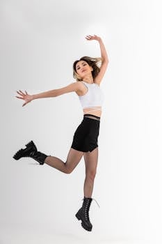 A stylish model in a white top and black shorts jumping energetically in a studio setting.