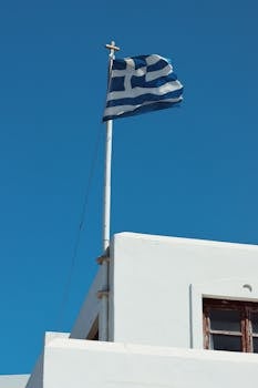 Greek flag waving on a white building under a clear blue sky in Naxos, Greece.
