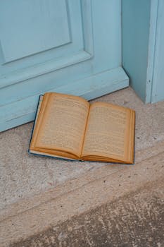 An open vintage book lies on a rustic wooden floor next to a blue door.
