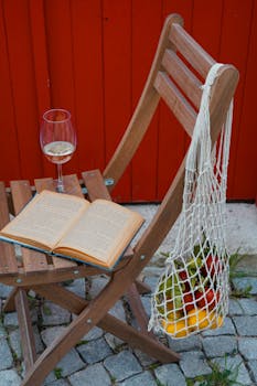 Relaxing outdoor setup with a book, glass of wine, and fruit on a wooden chair.