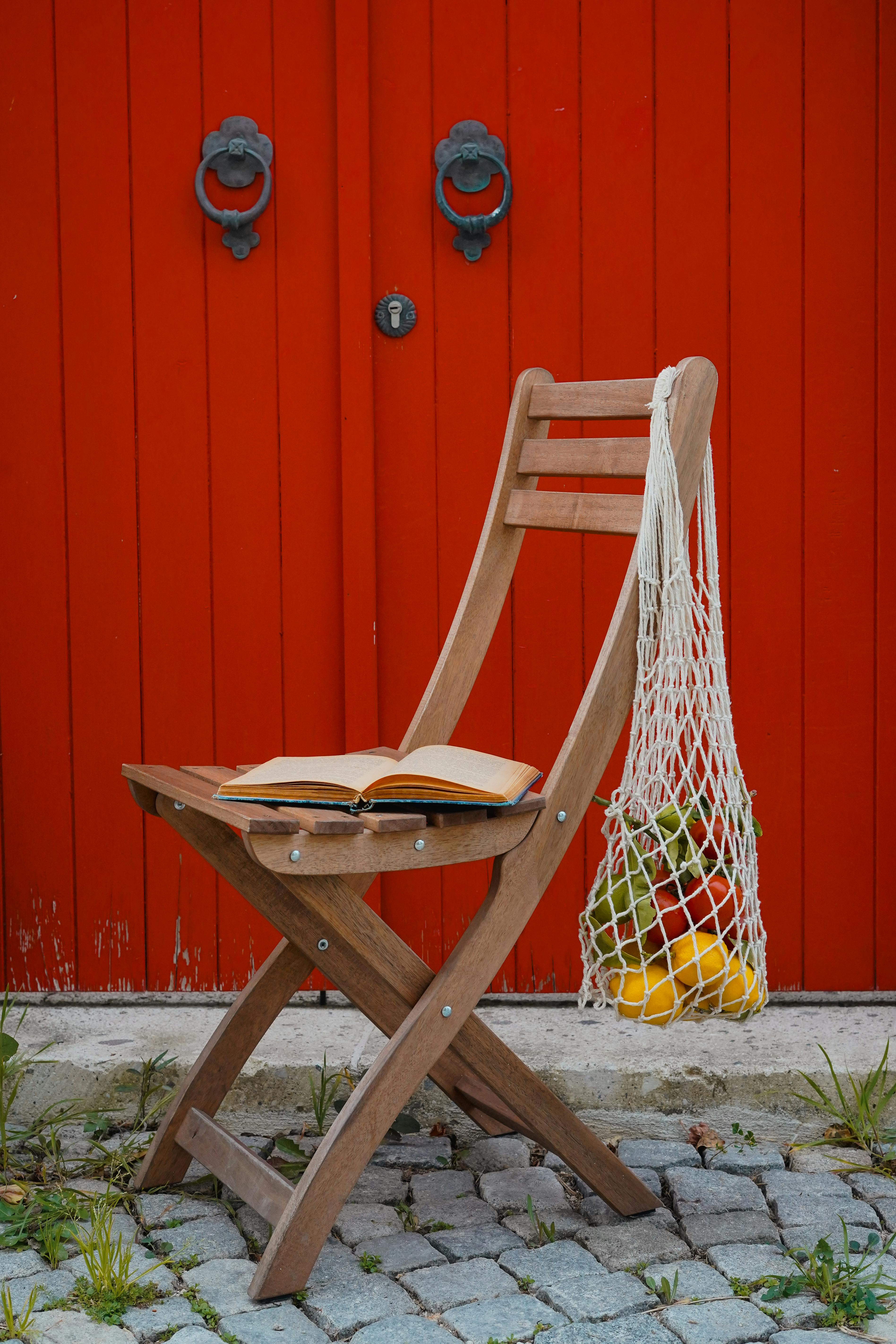 Wooden chair with book and net bag of fruit by red door. Rustic charm.