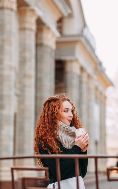 Stylish young woman with red hair enjoys a coffee outdoors in a sunny urban setting.