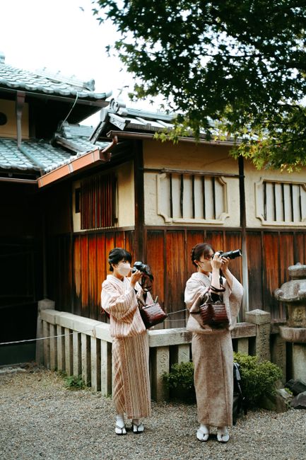Two women in kimono capturing moments with cameras outside a traditional Japanese building.