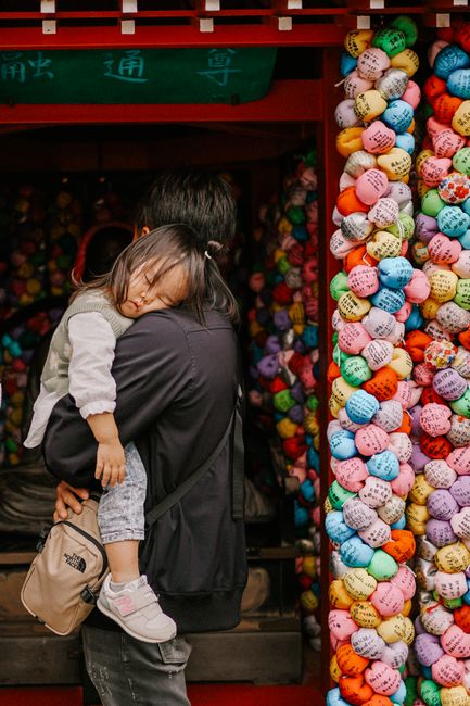 A father carries his sleeping child at a colorful Japanese temple adorned with prayer balls.