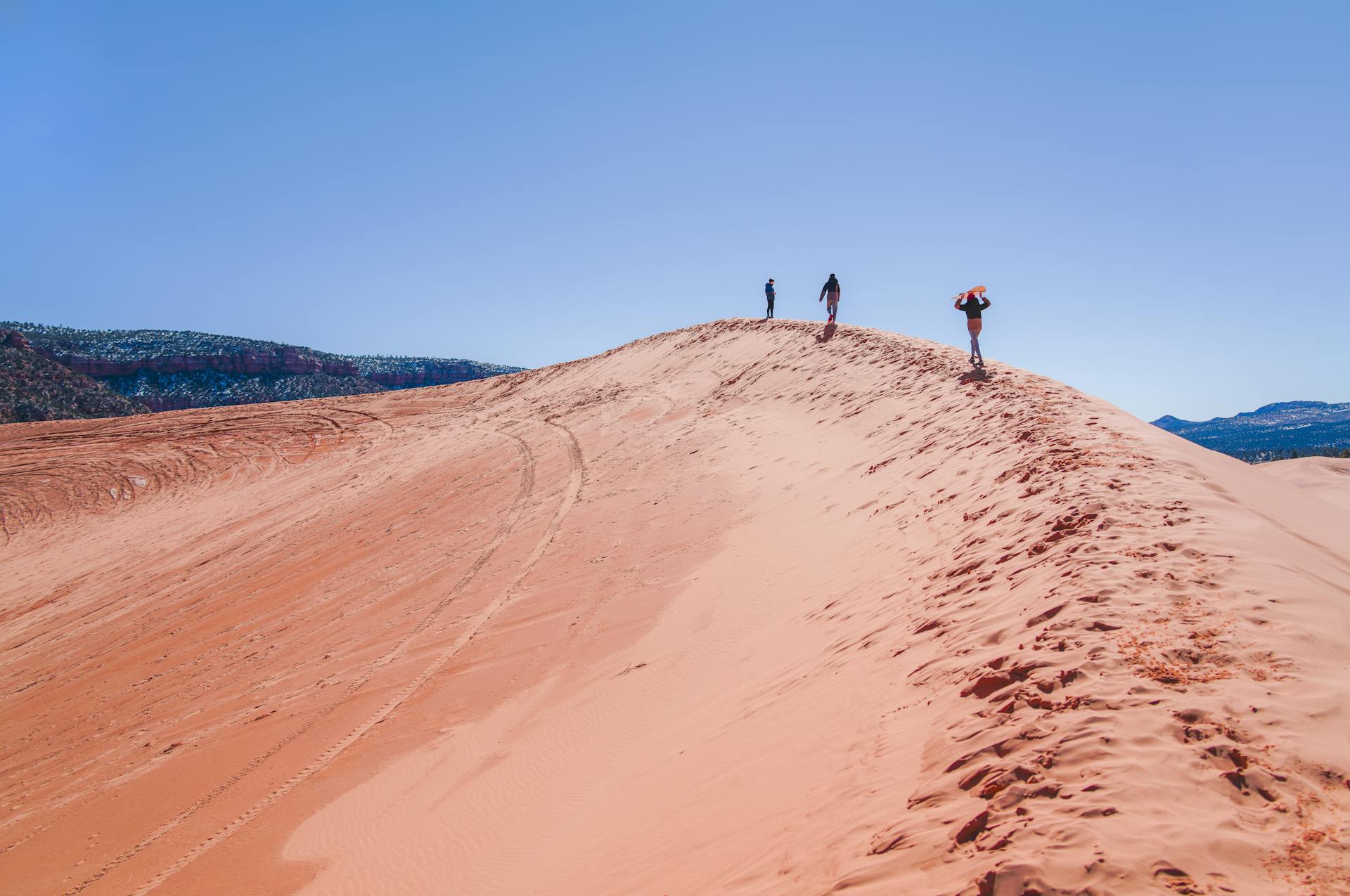 Three hikers atop the vibrant Coral Pink Sand Dunes in Utah under a clear blue sky.