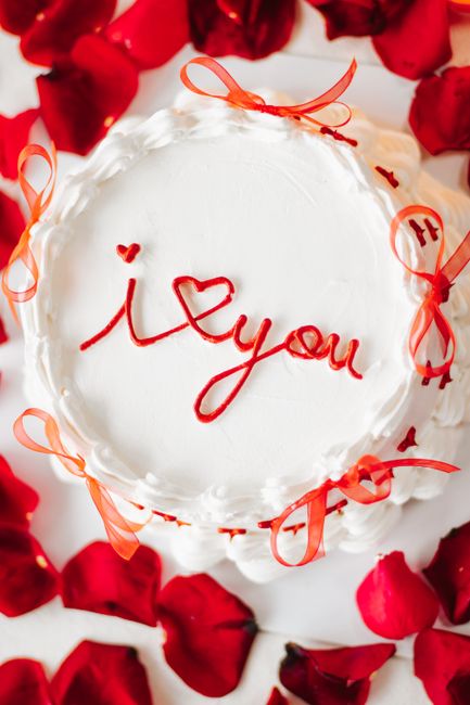 Top view of a romantic white cake with red 'I Love You' icing, surrounded by rose petals.