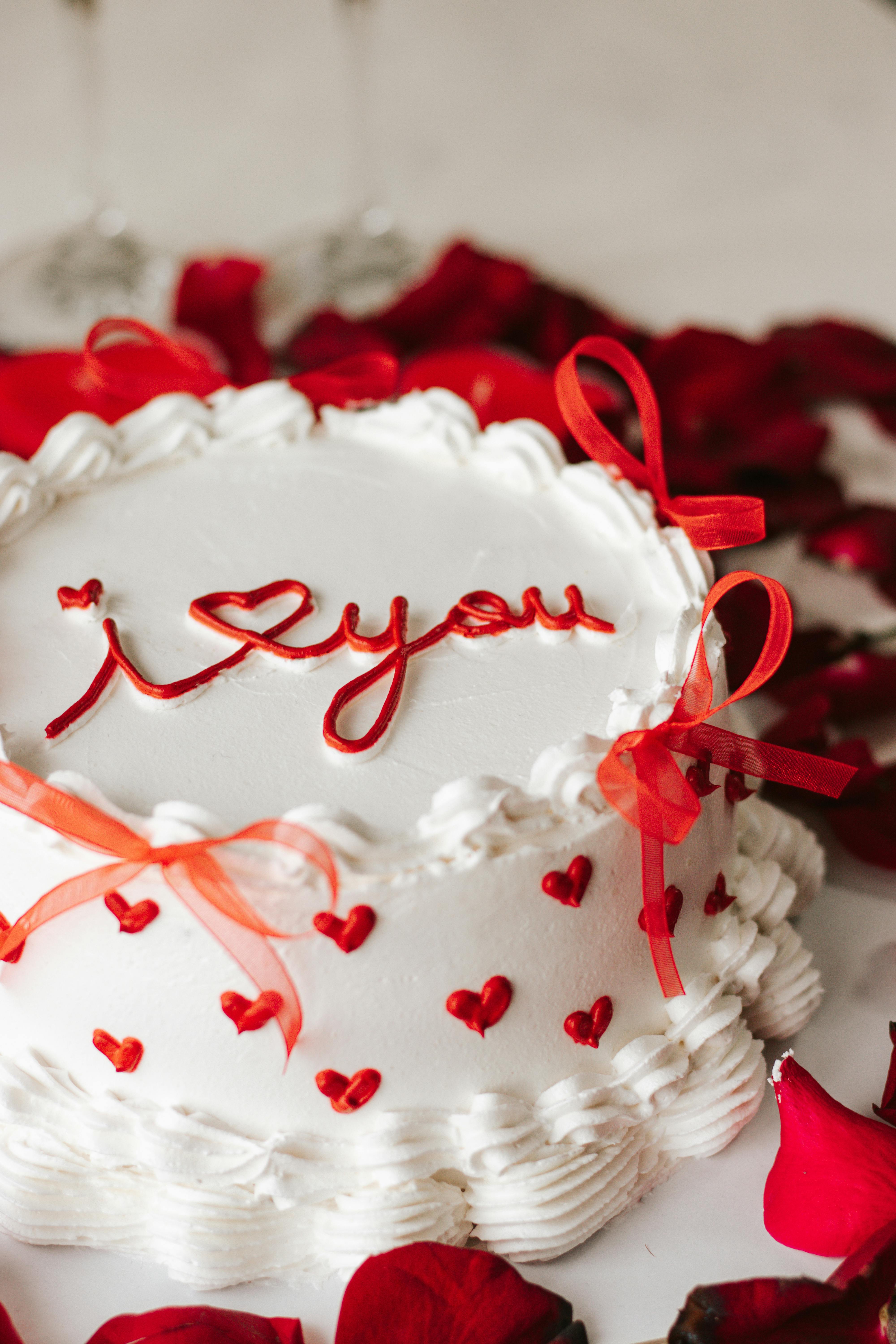 White cake with I love you icing, red bows, surrounded by rose petals, ideal for romantic occasions.