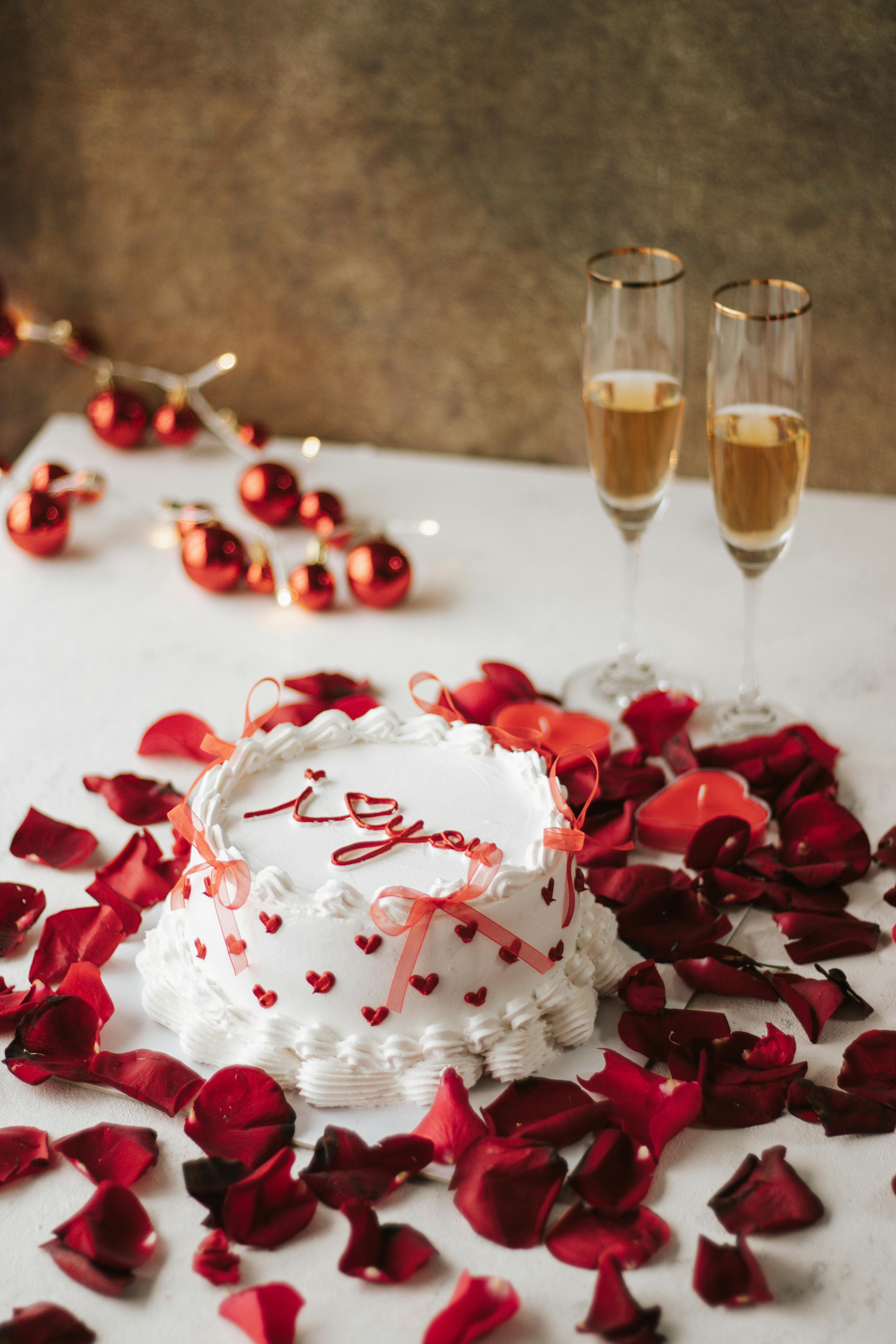 Elegant white cake with red hearts, rose petals, and champagne flutes on a romantic setting.