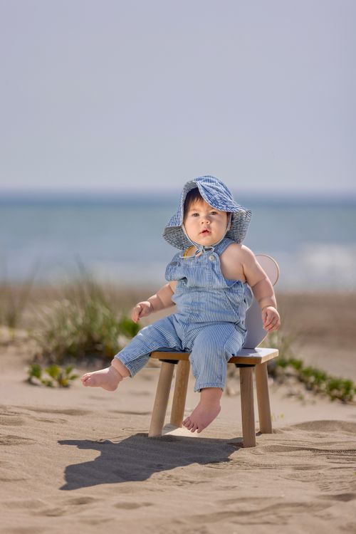 Free Cute baby in blue striped outfit sitting on a beach chair by the sea on a sunny day. Stock Photo