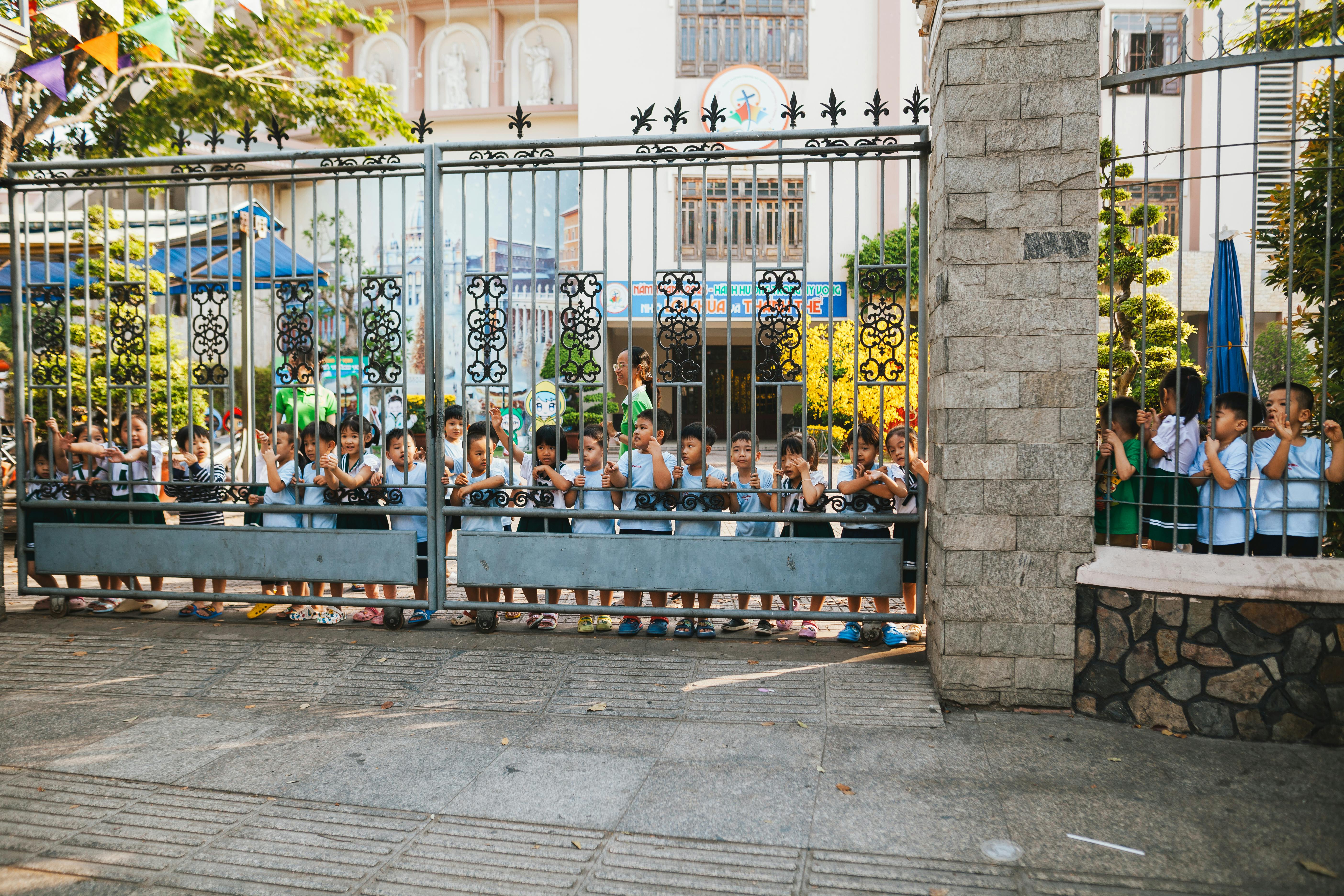 A group of young children, lined up and peering through a decorative school gate, enjoying a sunny day outdoors.