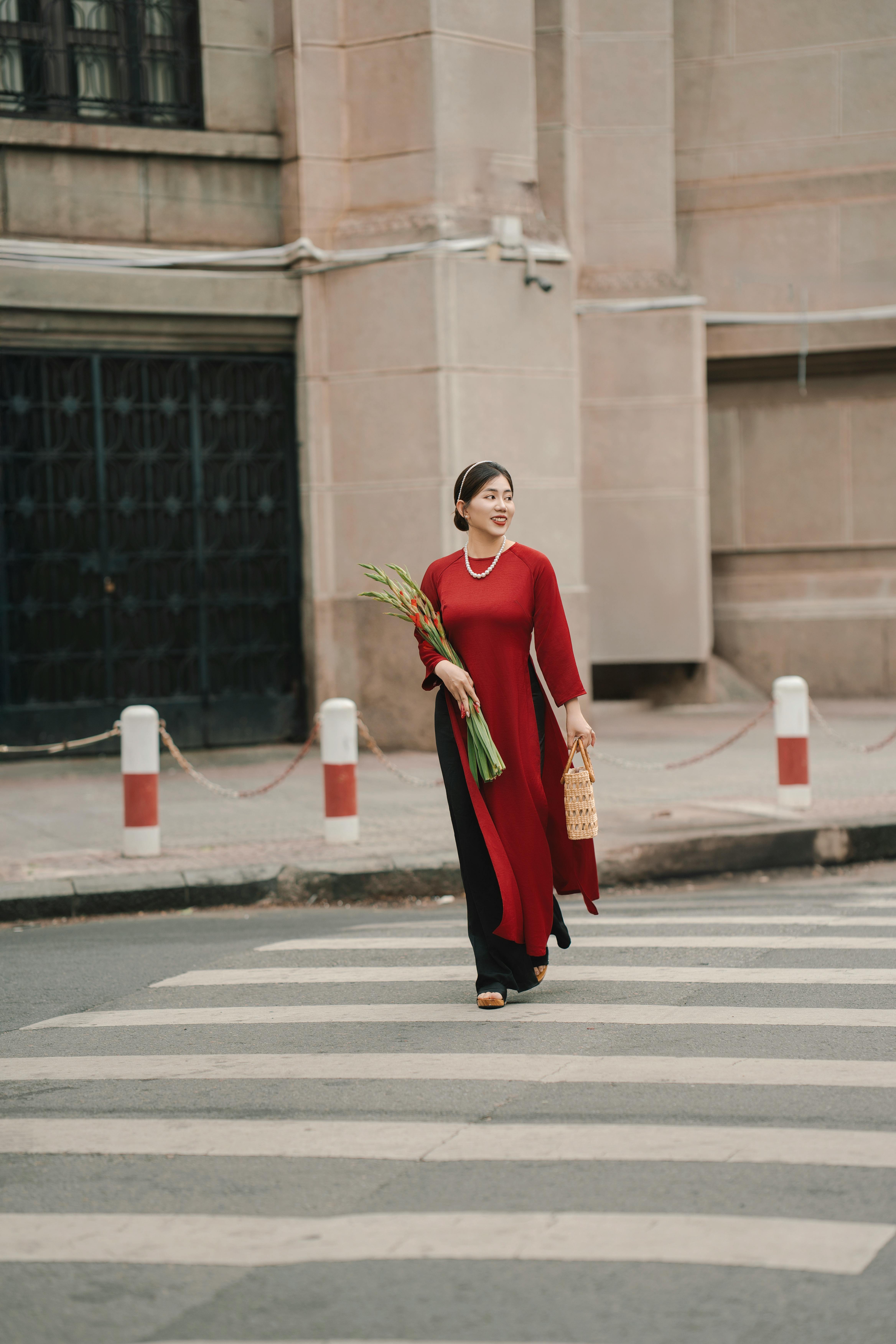 A woman in a red dress carrying flowers and a basket crosses a city street.