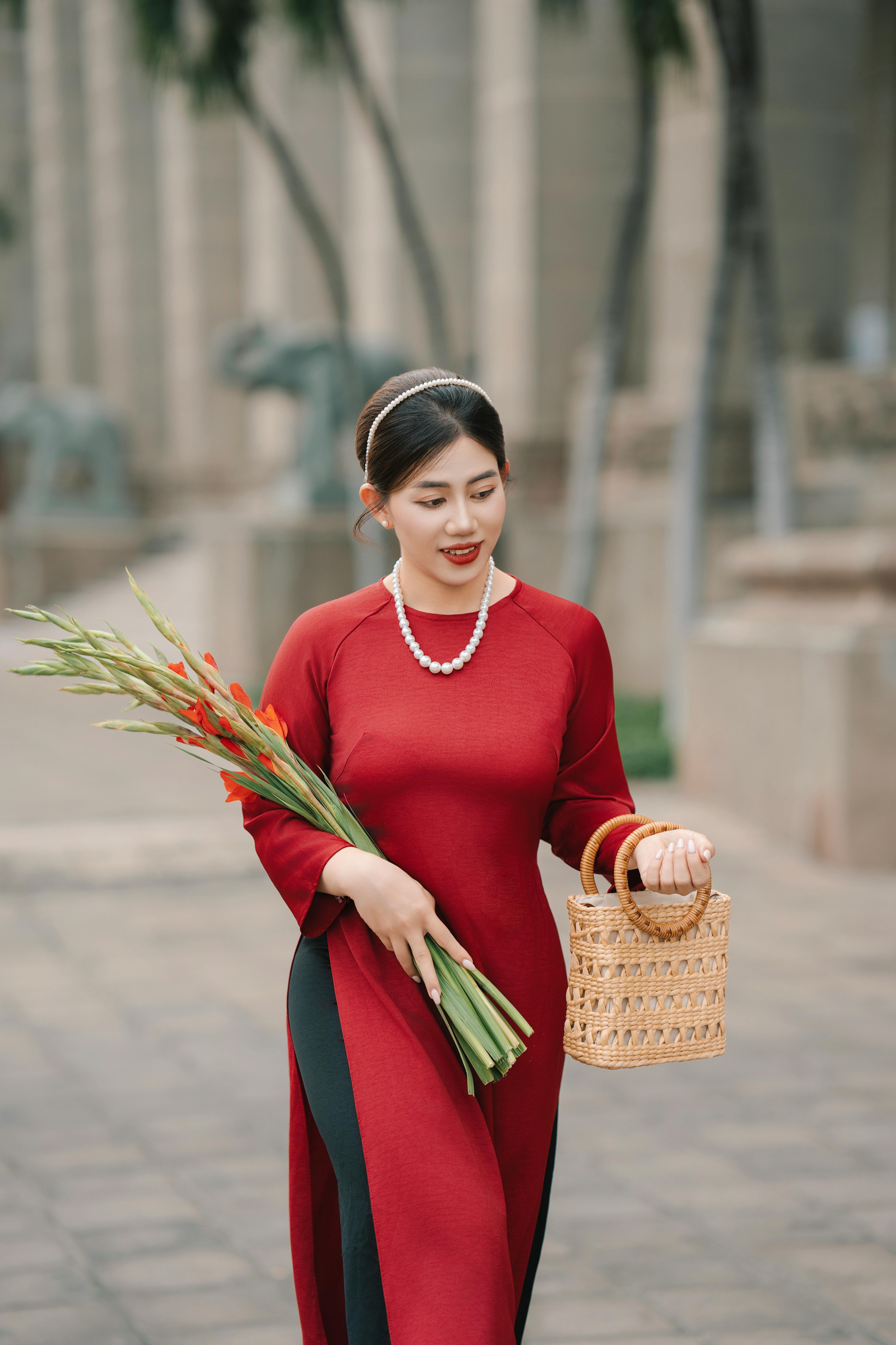 Elegant woman in a red dress carrying gladiolus flowers and a woven basket outdoors.