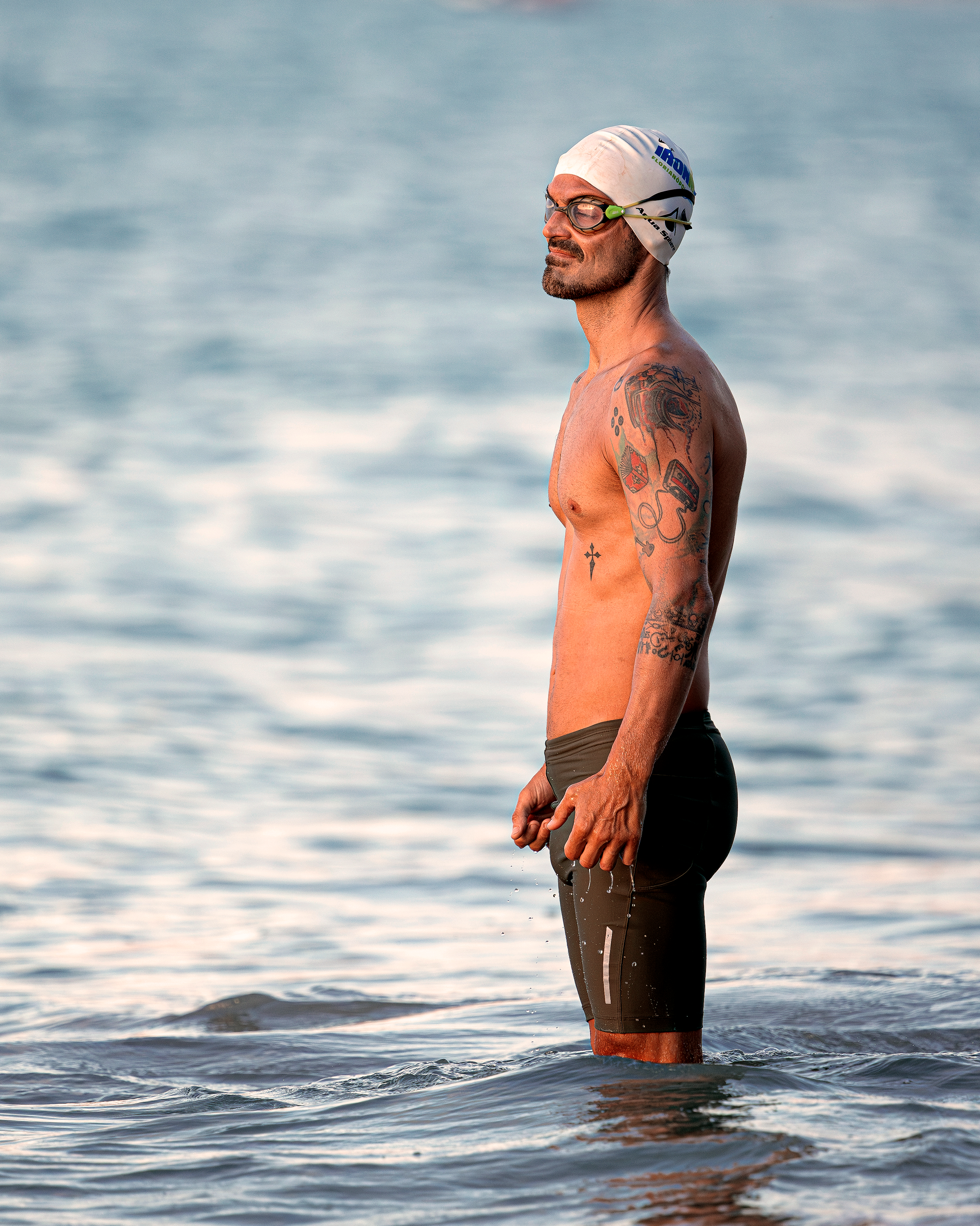 Tattooed male swimmer standing in ocean water wearing swim goggles and cap.
