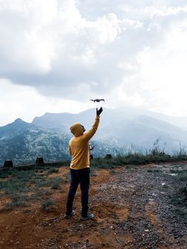 A person in a yellow hoodie launches a drone against a scenic mountain backdrop.