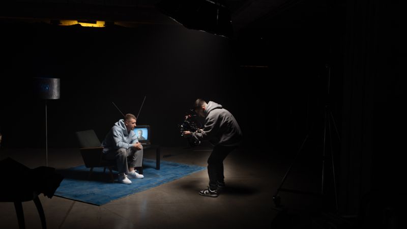 Film production shoot indoors with cinematographer filming an actor sitting, studio lighting.