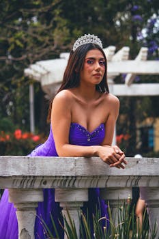 A young woman in a purple dress and tiara poses on a stone balcony in a park.