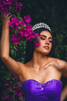 Elegant woman in fantasy cosplay with crown and purple dress posing among vibrant flowers.