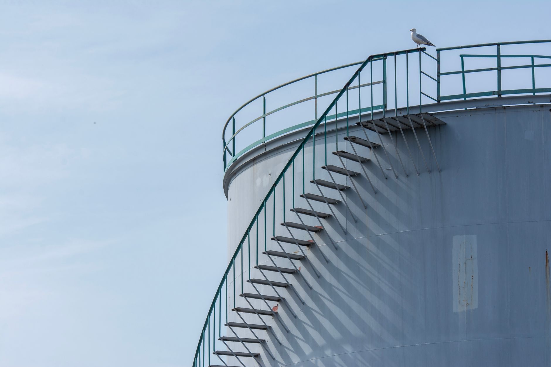 A seagull rests on the stairs of an industrial storage tank, casting shadows.