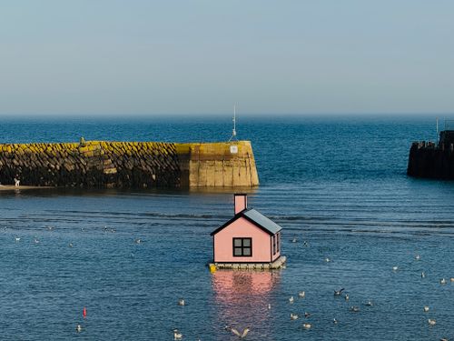 Free A pink house floats in a harbor amidst seagulls, with a stone pier and clear sky. Stock Photo