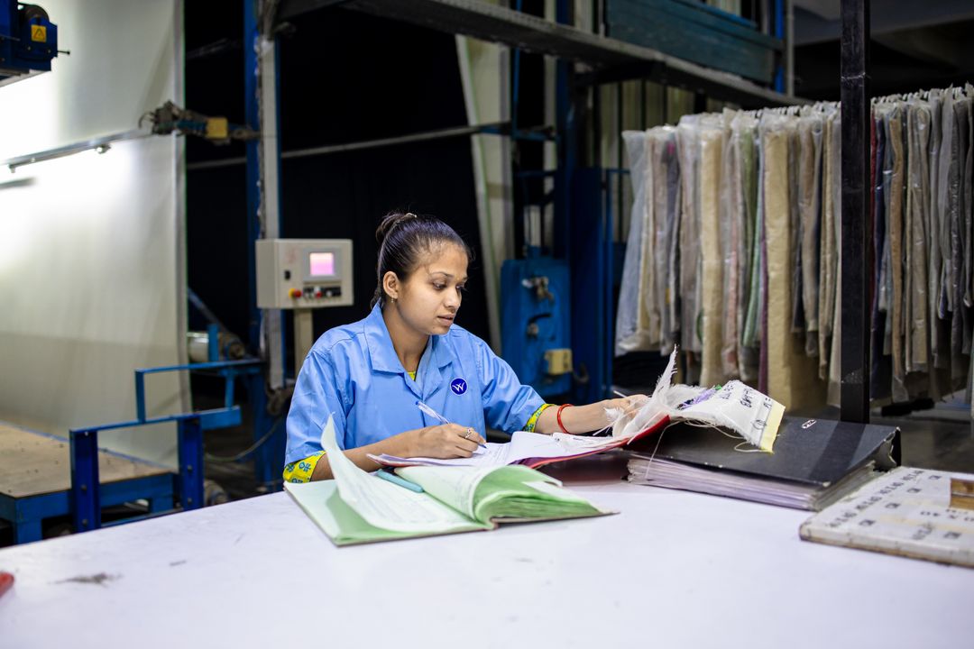 A female textile worker in a factory reviews documents at a workstation.