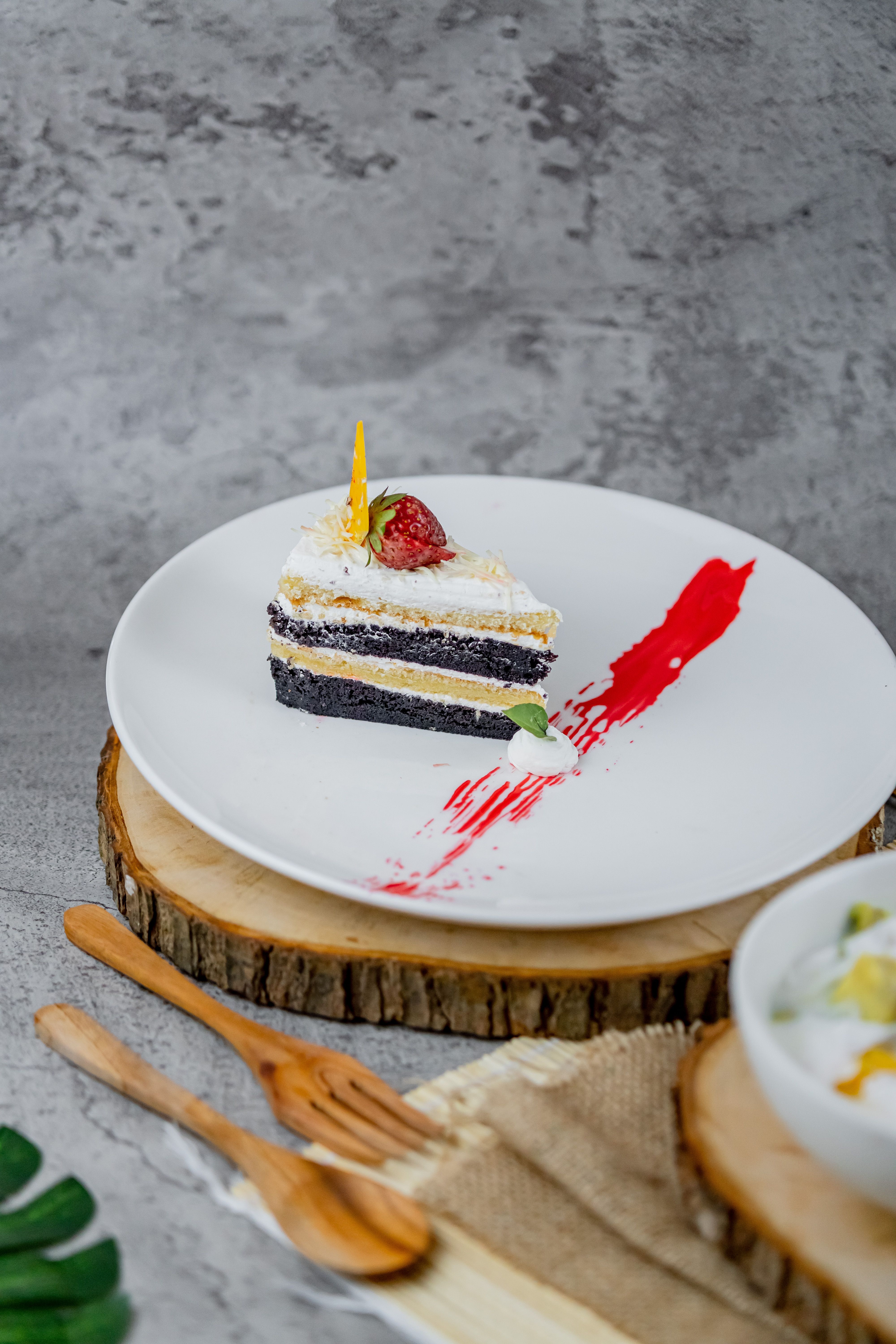 A slice of layered chocolate and vanilla cake garnished with strawberry, beautifully presented on a wooden slab.