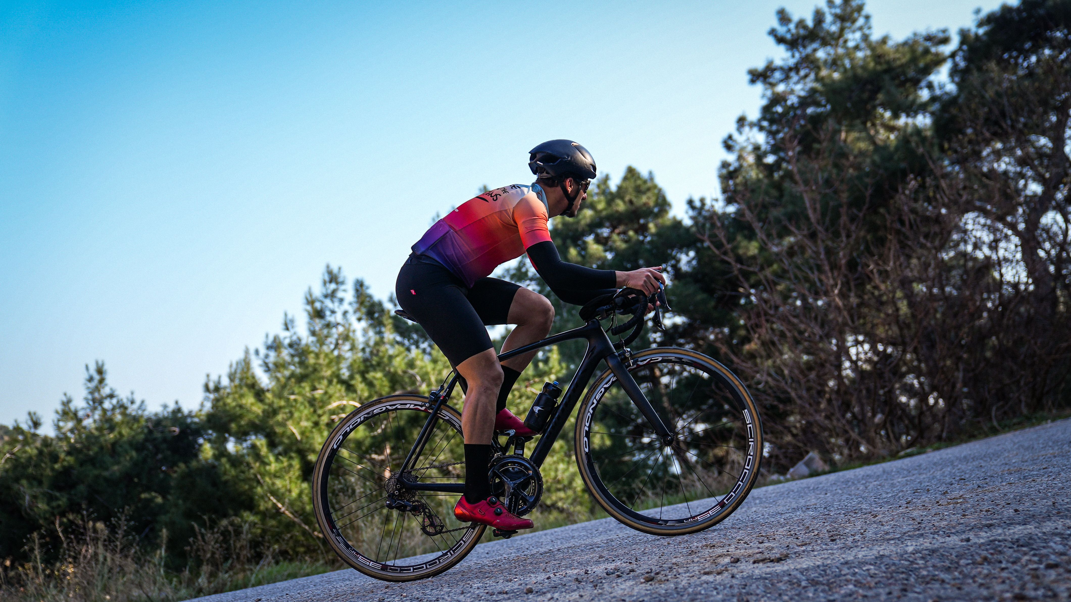 A cyclist wearing a vibrant jersey rides uphill on a mountain road surrounded by trees.