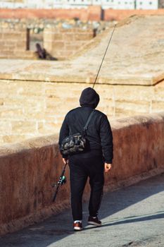 A solitary fisherman walks along a sandy urban path holding his fishing gear, showcasing outdoor adventure.