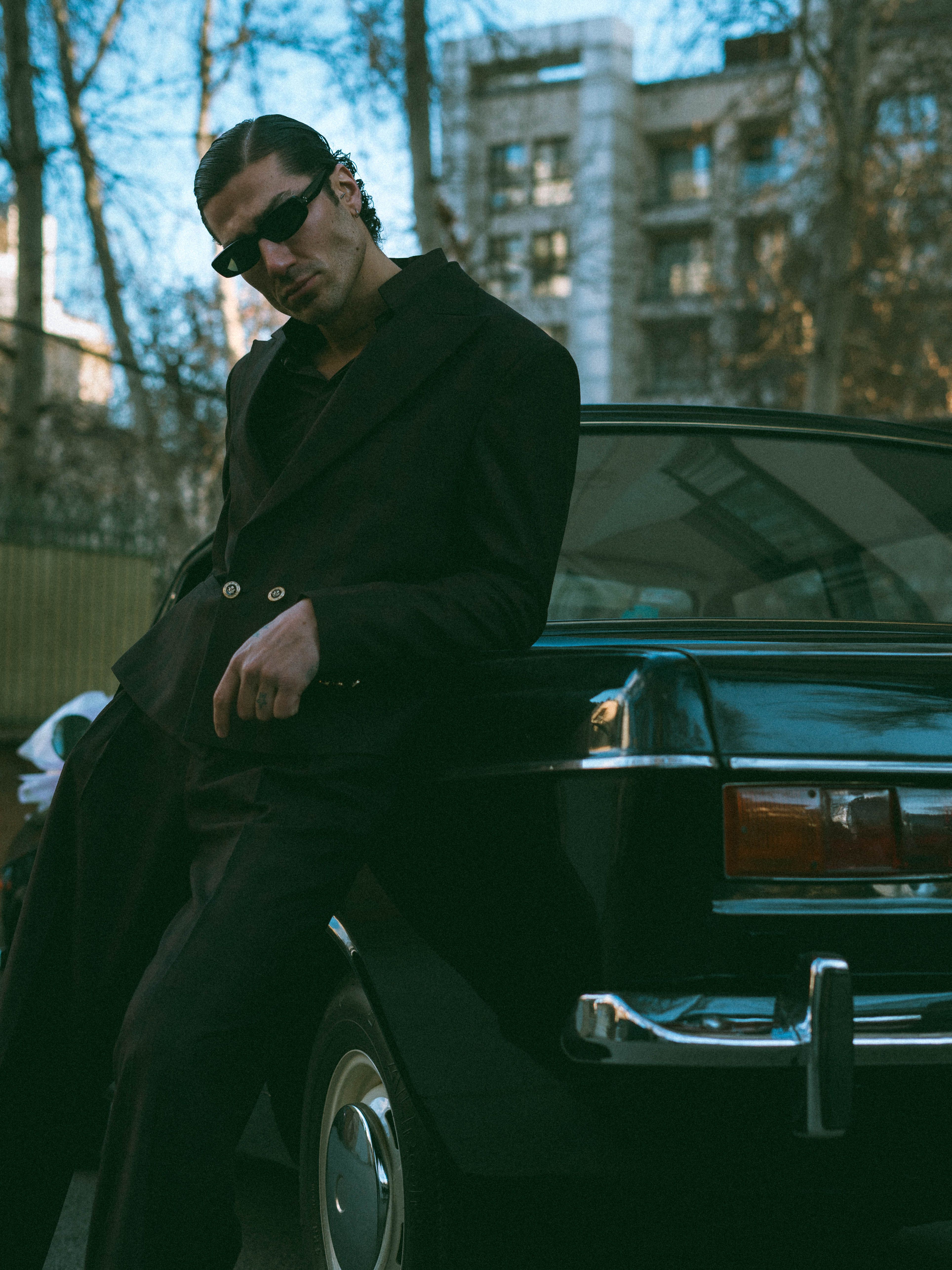 Man in sunglasses and suit leaning on vintage car in urban setting.